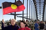 A couple are holding an Aboriginal flag as they walk on the Sydney Harbour Bridge, there is a crowd of people around them.