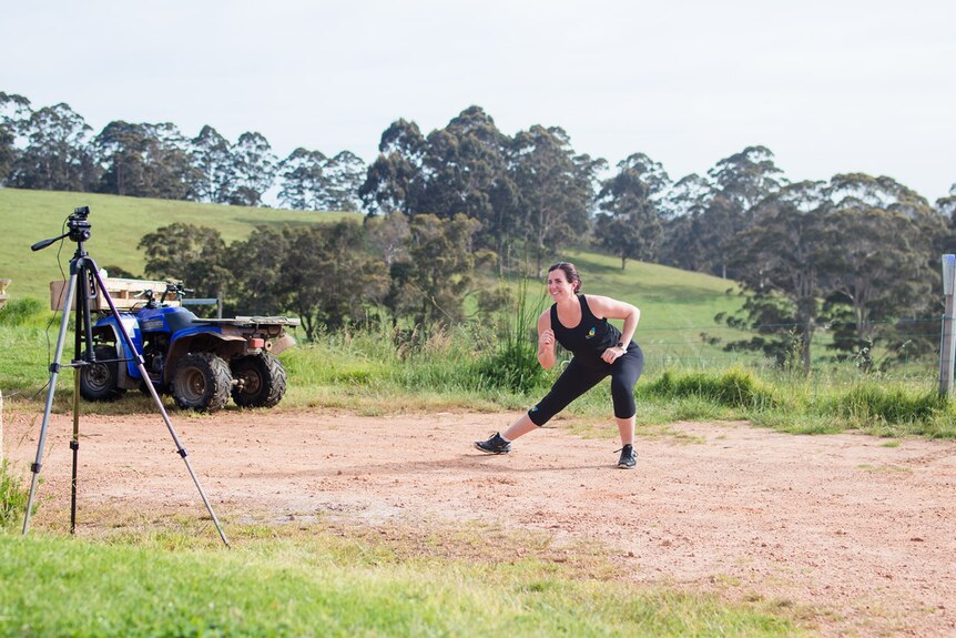 A woman in black lycra does a stretch in front of a video on a tripod, with a blue tractor in the background.