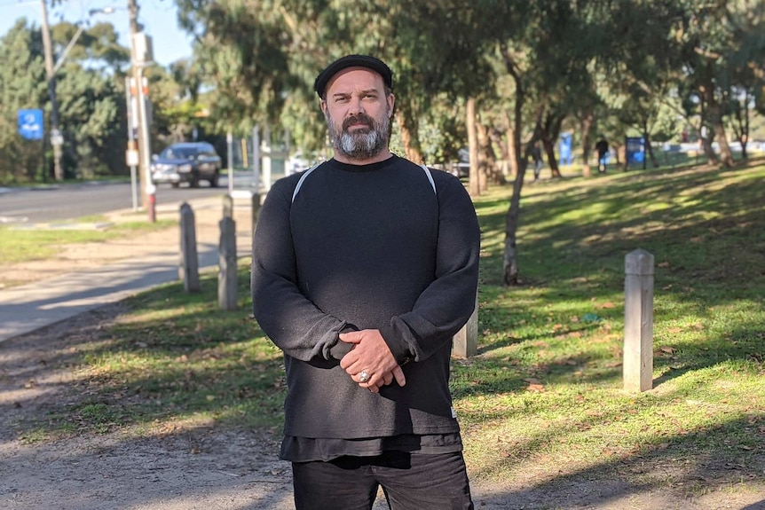 Maz Salt wears a black hat and jumper with hands crossed in front of him standing in a park on a sunny day.