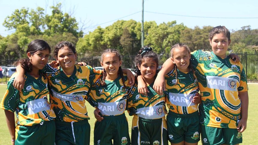 A group of young girls smile in green patterned jerseys with their arms around each other on an oval