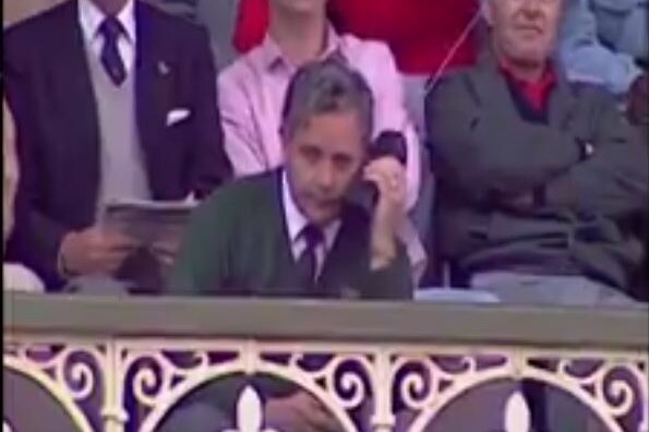 Grainy old TV footage of a man in a green blazer sitting among the crowd at the SCG, listening into a large black mobile phone.