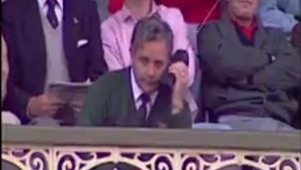 Grainy old TV footage of a man in a green blazer sitting among the crowd at the SCG, listening into a large black mobile phone.