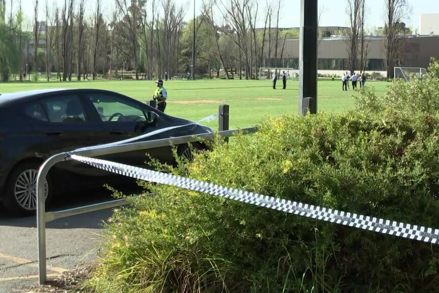 A car parked next to a football pitch. Police tape is stretched across part of the area. Several police are on the pitch.