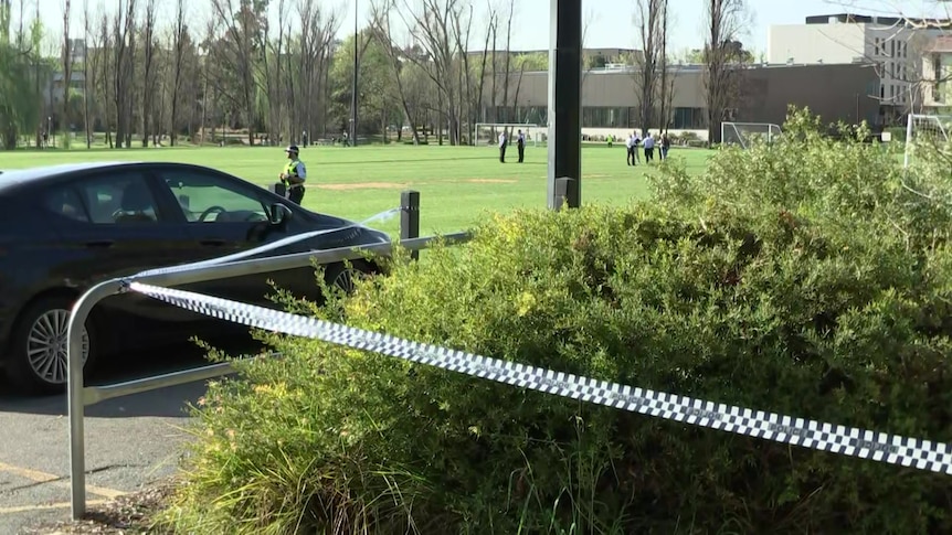 A car parked next to a football pitch. Police tape is stretched across part of the area. Several police are on the pitch.