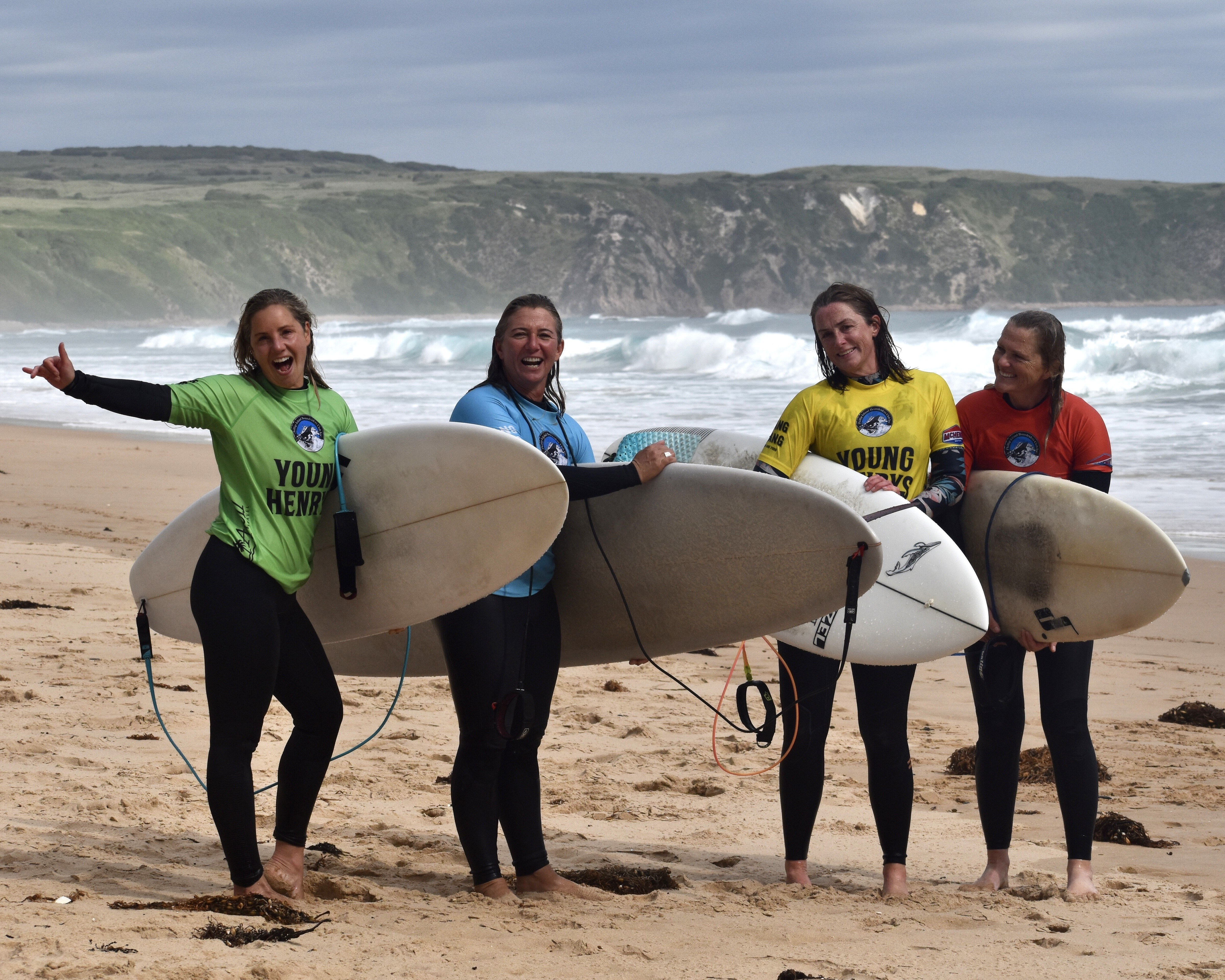 Four women stand on the beach holding surfboards wearing different coloured rash vests.