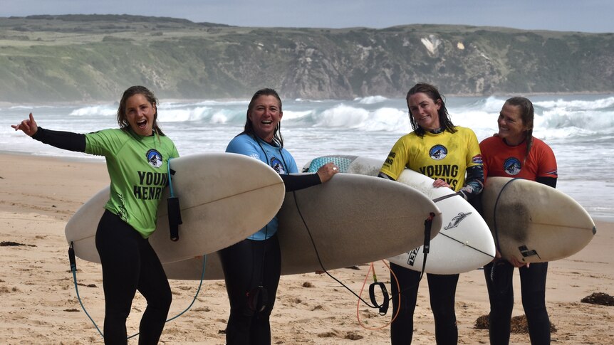 Four women stand on the beach holding surfboards wearing different coloured rash vests.