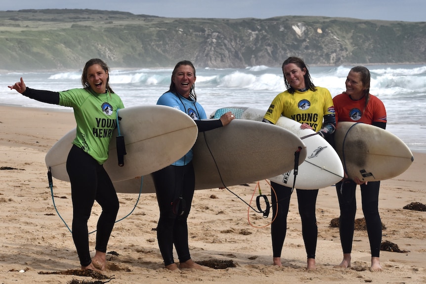 Four women stand on the beach holding surfboards wearing different coloured rash vests.