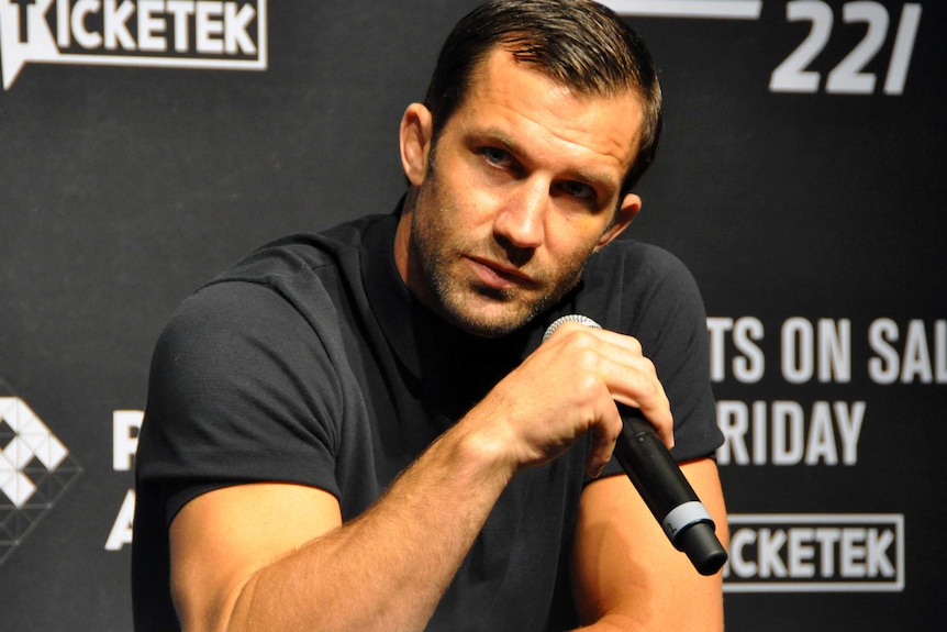 A UFC fighter wearing a black T-shirt holds a microhone while speaking at a press conference.
