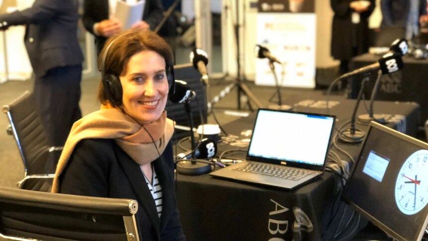 A woman with headphones on faces camera with mic, laptop, and radio broadcast equipment behind.