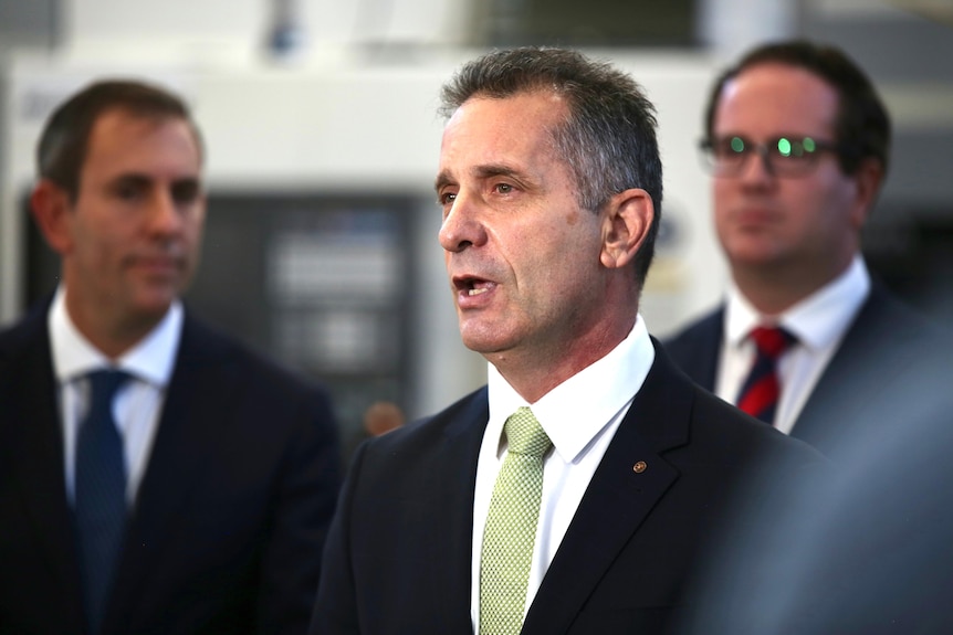 A man in a suit speaks at a press conference watched on by two other men in suits. 