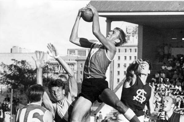 An Australian rules player leaps high into a pack and grasps the ball in both hands as players grimace around him.