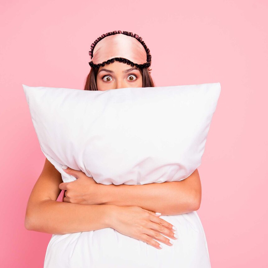 A woman hugging a pillow and wearing a sleeping mask on her head with an astonished look on her face