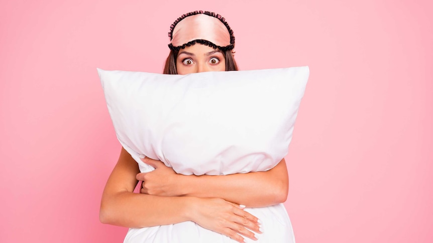 A woman hugging a pillow and wearing a sleeping mask on her head with an astonished look on her face