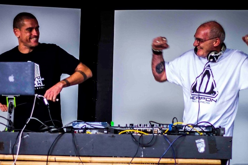 Photo of two men standing behind DJ decks smiling at each other.