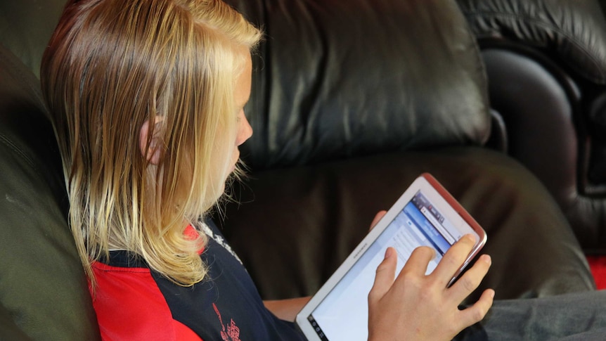 A blond child whose face is turned away from the camera sits on a couch and uses an iPad.
