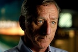 Peter Charley looks to an interviewer sitting beside the camera. Blurred coloured lights fill the background.