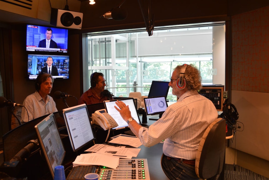 Jon Faine in studio interviewing John Paul Young with four computer monitors and two TV screens in front of him.