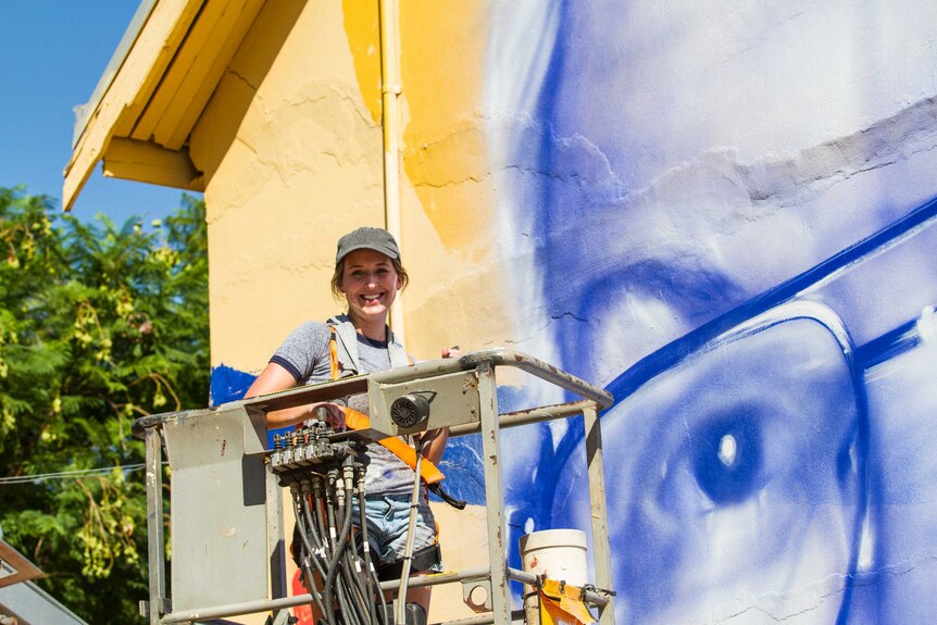 A woman wearing a cap on a cherry picker, painting a mural on a building.