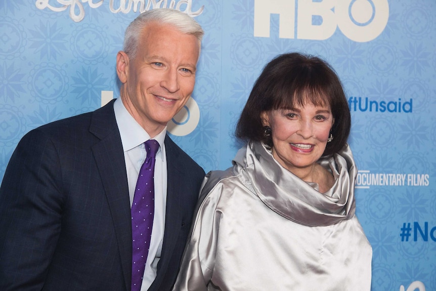 Gloria Vanderbilt wearing a silver gown and standing next to Anderson Cooper in front of a press wall