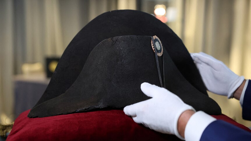 A pair of hands in white gloves tentatively place Napoleon's hat on a red cushion. 
