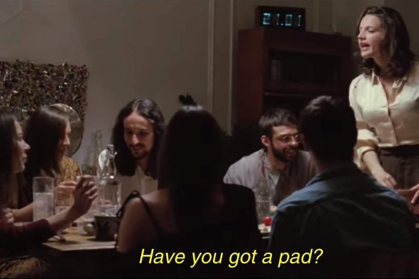 A woman standing up at a dinner speak to someone at the other end of the table and the caption read, "Have you got a pad?"