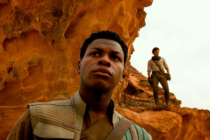 John Boyega looks into the distance while a man behind him stands on a red rock formation.