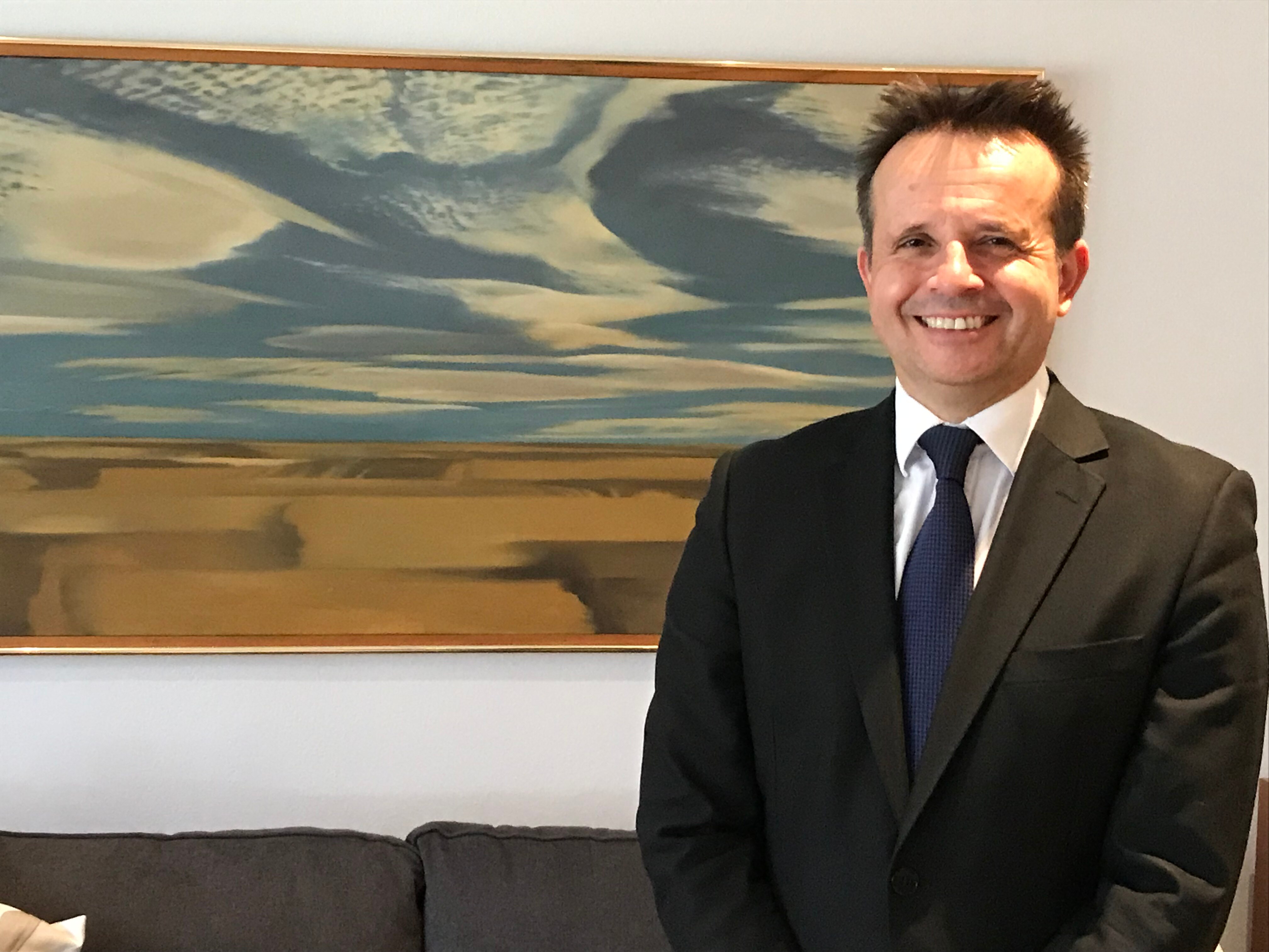 A man wearing a black suit and navy tie with brown hair smiling in front of a painting. 
