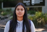 A woman of Indian descent stands looking solemn on a university campus.