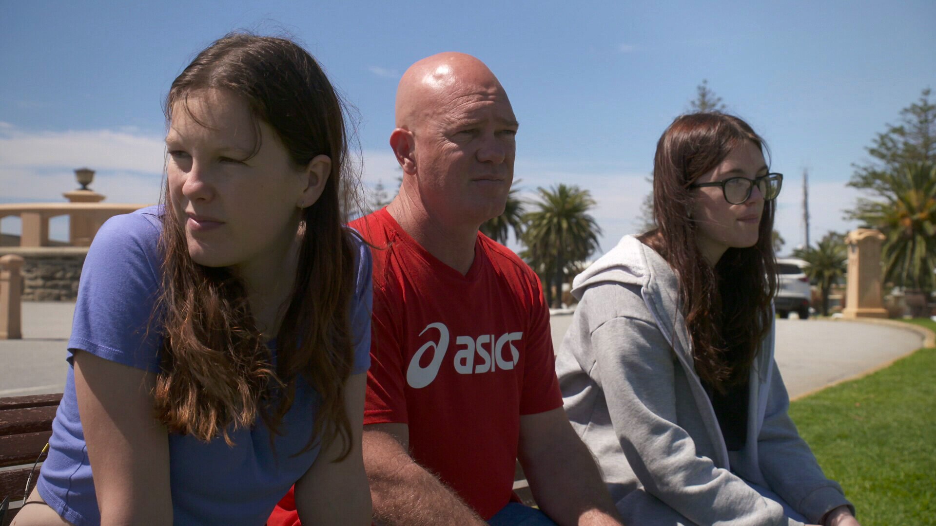 Bald man in red Asics T-shirt sitting down next to two teen girls with long brown hair