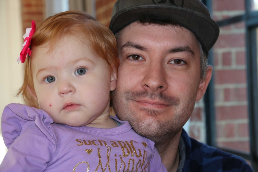 A young toddler with red hair is held by her father, a man wearing a cap.