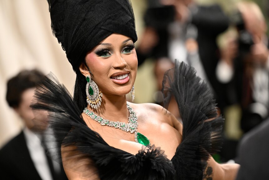 Cardi B wearing a black tulle off-the-shoulder dress with a green gem necklace, matching earrings and a black headpiece.