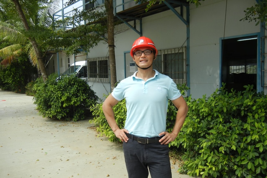 Yanging Tang smiles for the camera with his hands on his hips, wearing a bright red hard hat in front of low building.