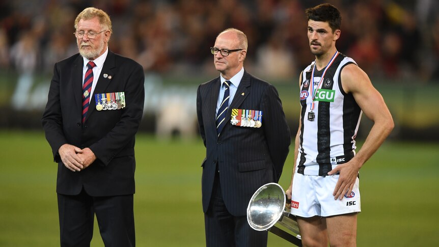 Scott Pendlebury stands holding a trophy at his side next to two men in suits with medals on their jackets.