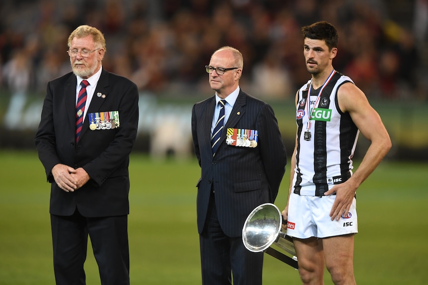 Scott Pendlebury stands holding a trophy at his side next to two men in suits with medals on their jackets.