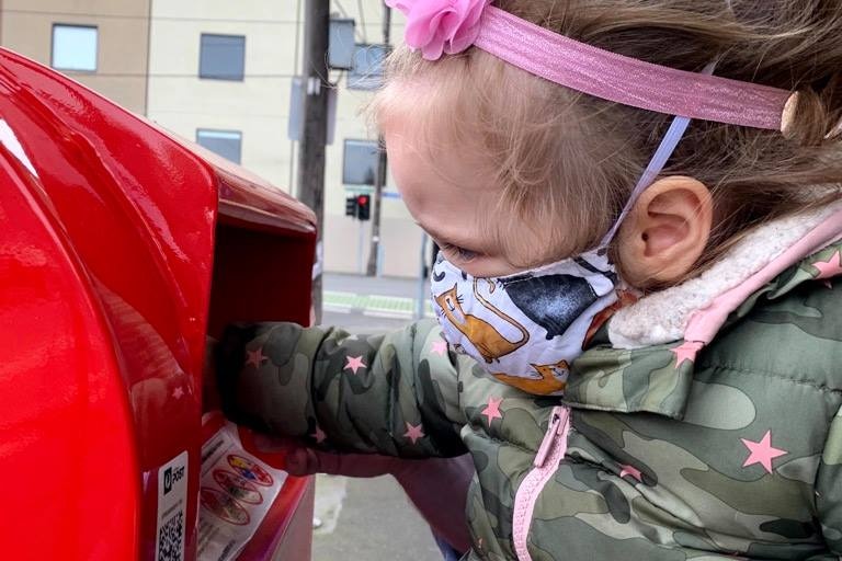 A small girl wearing a mask puts mail in a post box.