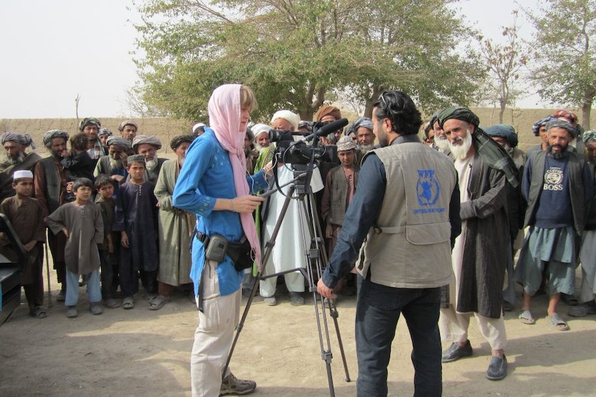 Woman wearing a scarf, operating a camera and interviewing a man as a crowd of men and boys watch.