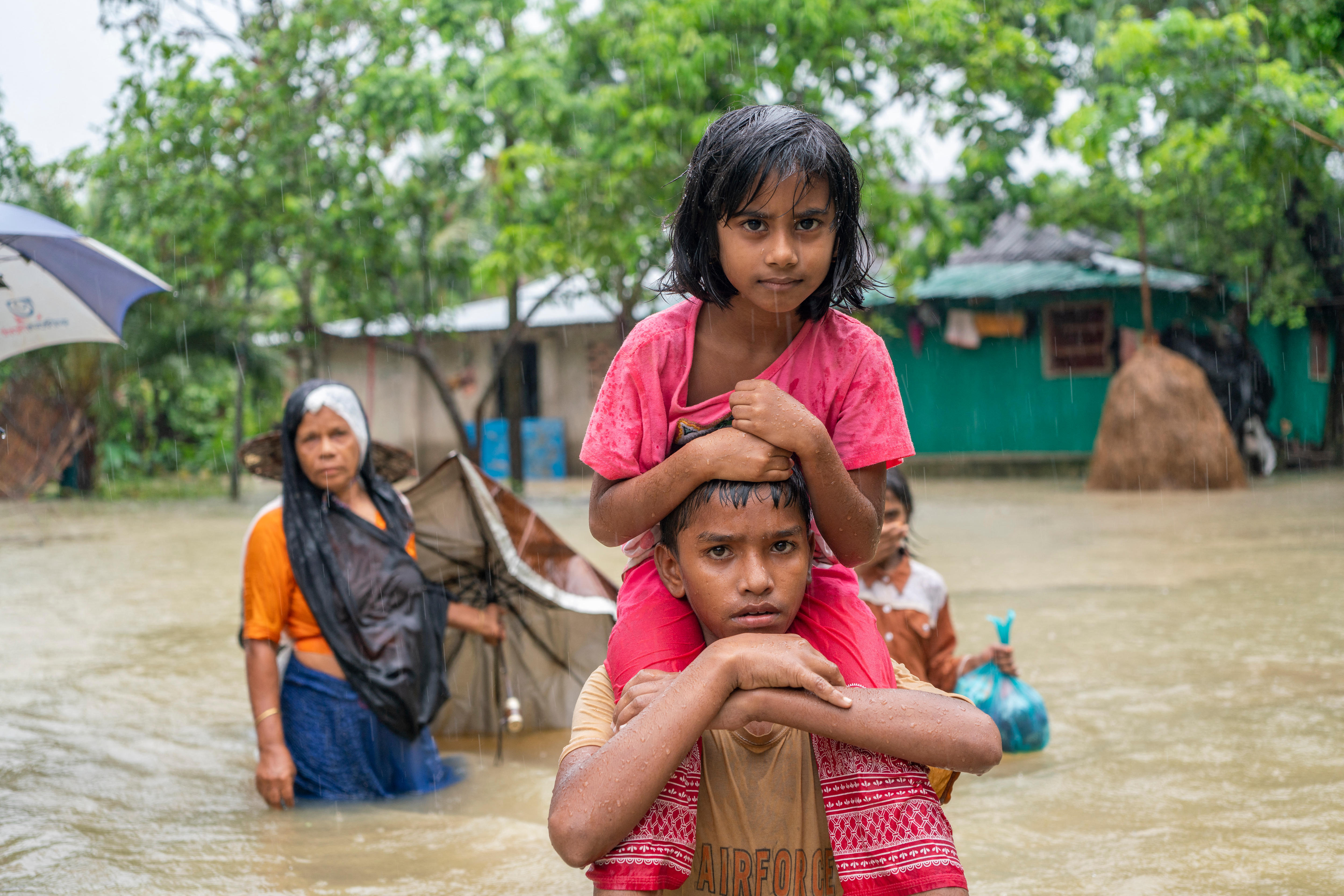 A close-up shot of a young boy carrying a girl on his shoulders as they navigate through deep floodwaters 