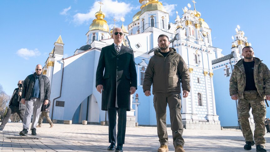 Biden walks next to Zelenskyy, behind them a glinting white and gold cathedral and a deep blue sky