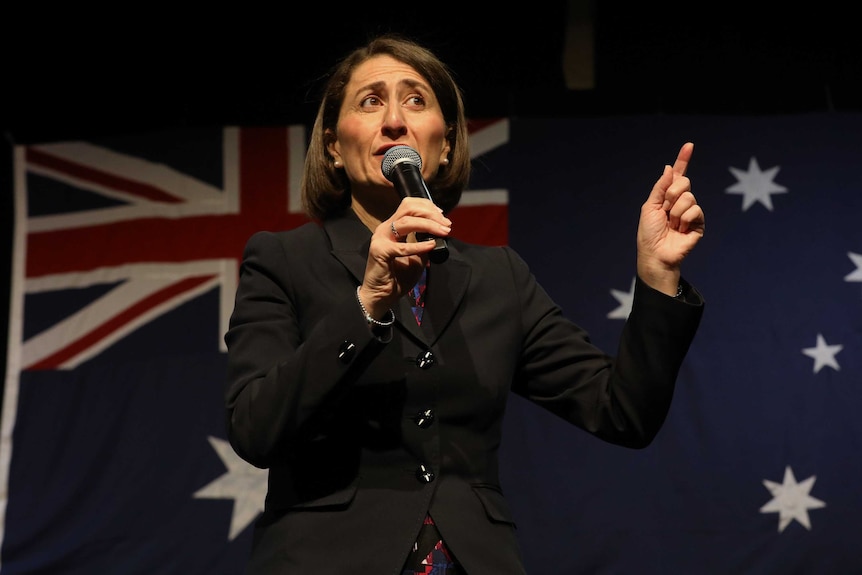 The premier talks into a microphone and points with her other hand. She's standing in front of an Australian flag