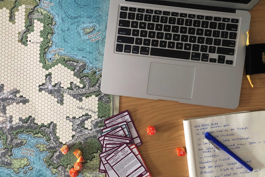 A map for a Dungeons and Dragons game, a laptop, some dice, cards with spells and a notebook with some notes sit on a table.
