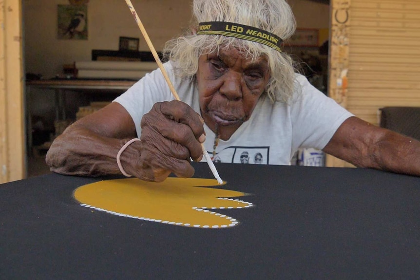 Artist wearing headband painting yellow image on black canvas.