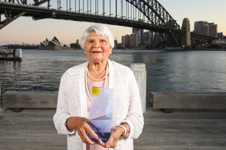 Older woman in a white top smiling in front of a bridge over water