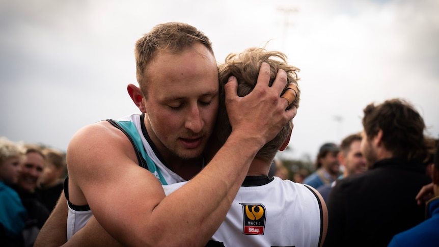 Two Australian Rules football players wearing white and blue guernseys embrace after winning a match