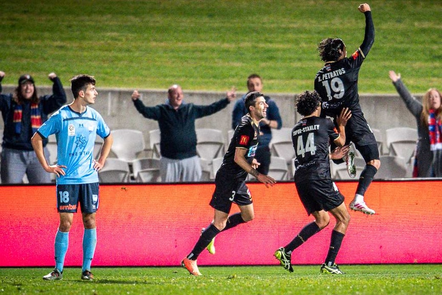 Newcastle Jets players jump and celebrate in front of their fans as Sydney FC players look disappointed.