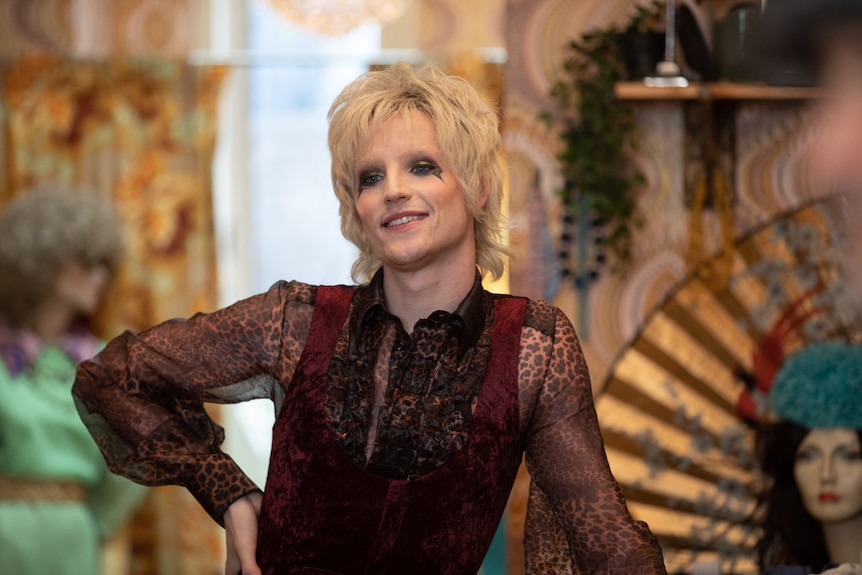 Film still of John McCrea as Artie with feathered hair and dark eyeshadow, standing in his vintage clothing store in Cruella