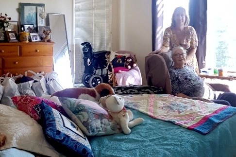 An elderly woman sits in a chair in a bedroom, with a younger woman standing beside her.