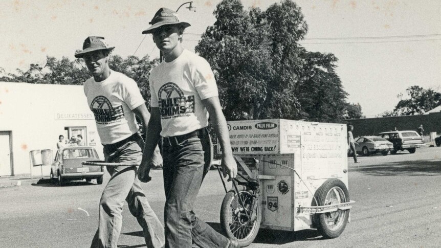 Black and white image of David Howison, smiling, and his son Mark, serious, wearing jeans, t-shirts and hats, with their cart.