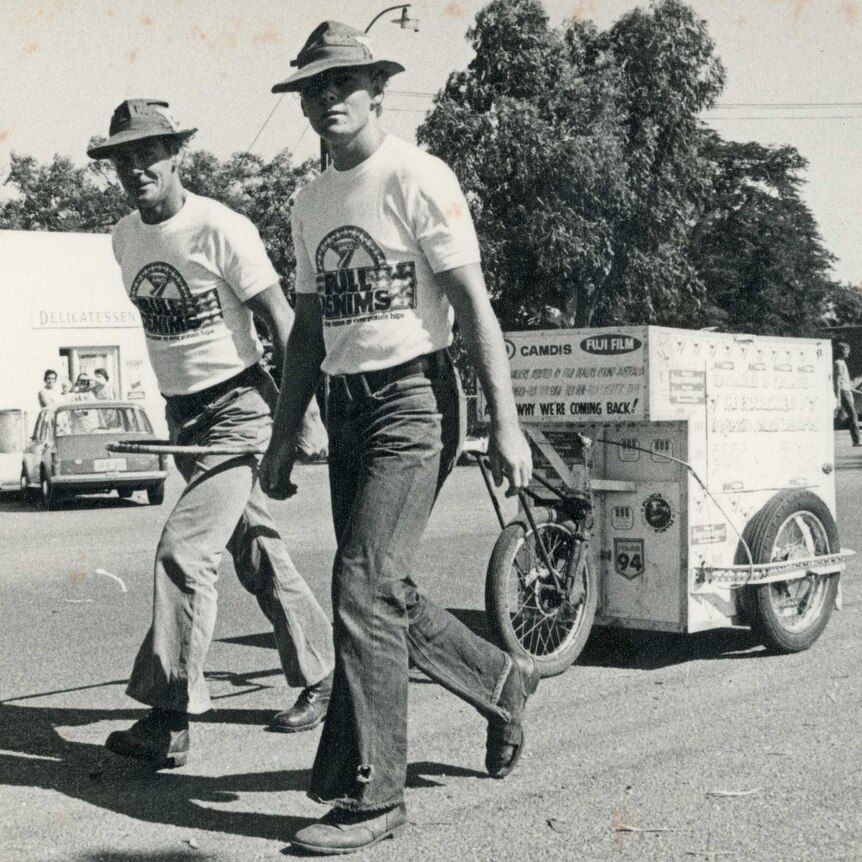Black and white image of David Howison, smiling, and his son Mark, serious, wearing jeans, t-shirts and hats, with their cart.