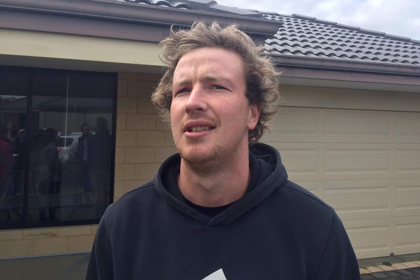 A head and shoulders shot of a man with blonde hair wearing a hoodie standing in front of a house.