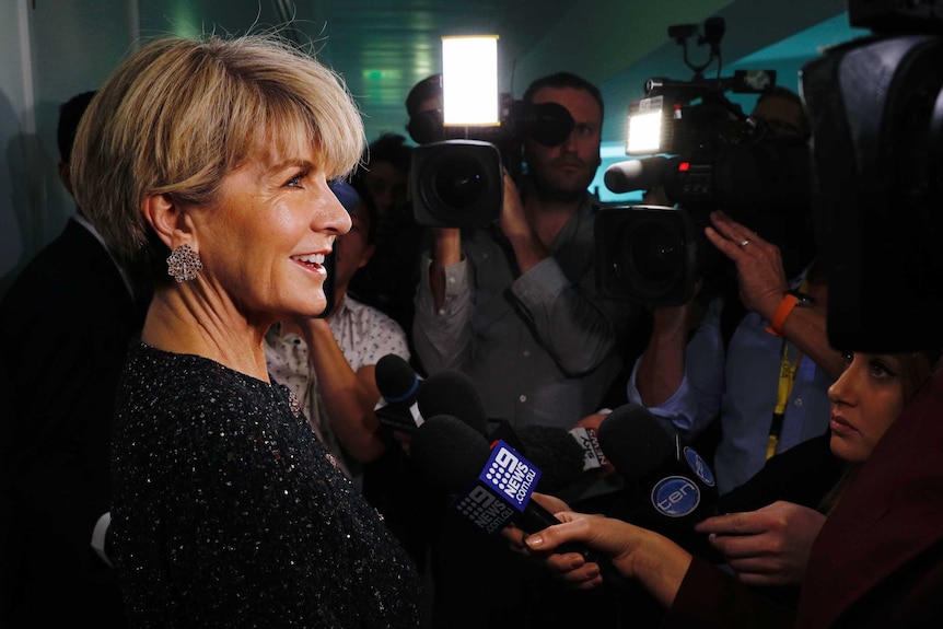 Julie Bishop, in a sequinned top and dazzlingly sparkly earrings, is surrounded by journalists with cameras and microphones.
